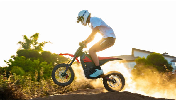 A person riding a red and black electric dirt bike on a dirt mound at sunset, with the rider wearing a white helmet, white t-shirt, and light blue sneakers kicking up dust in an outdoor setting.